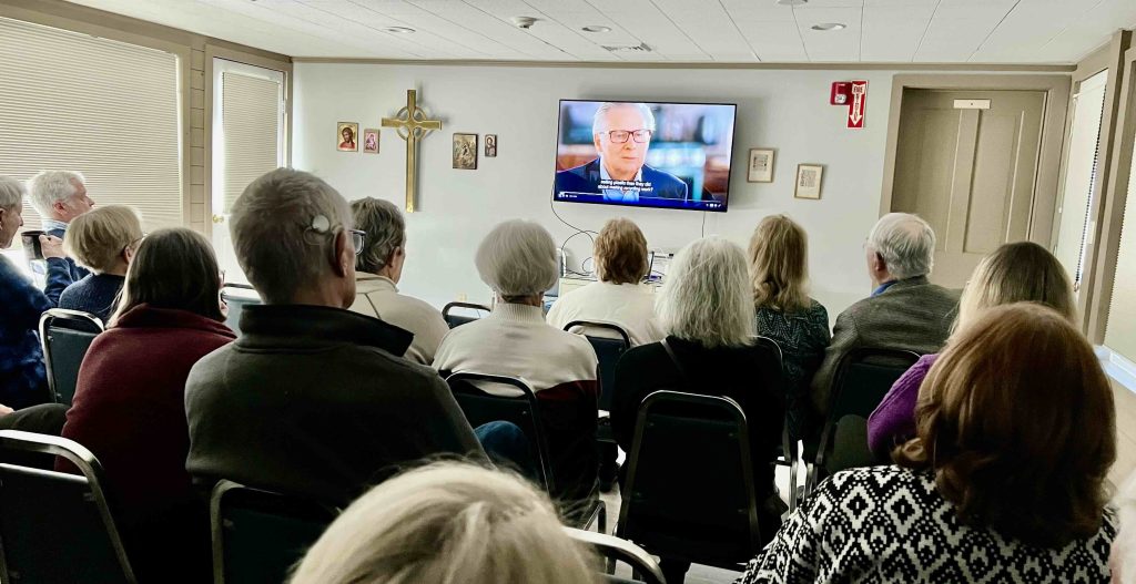a group of people sitting on black chairs watching a movie on a mounted screen