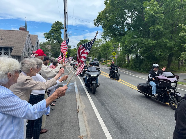 People standing along roadway holding flags as motorcycles are driving by.