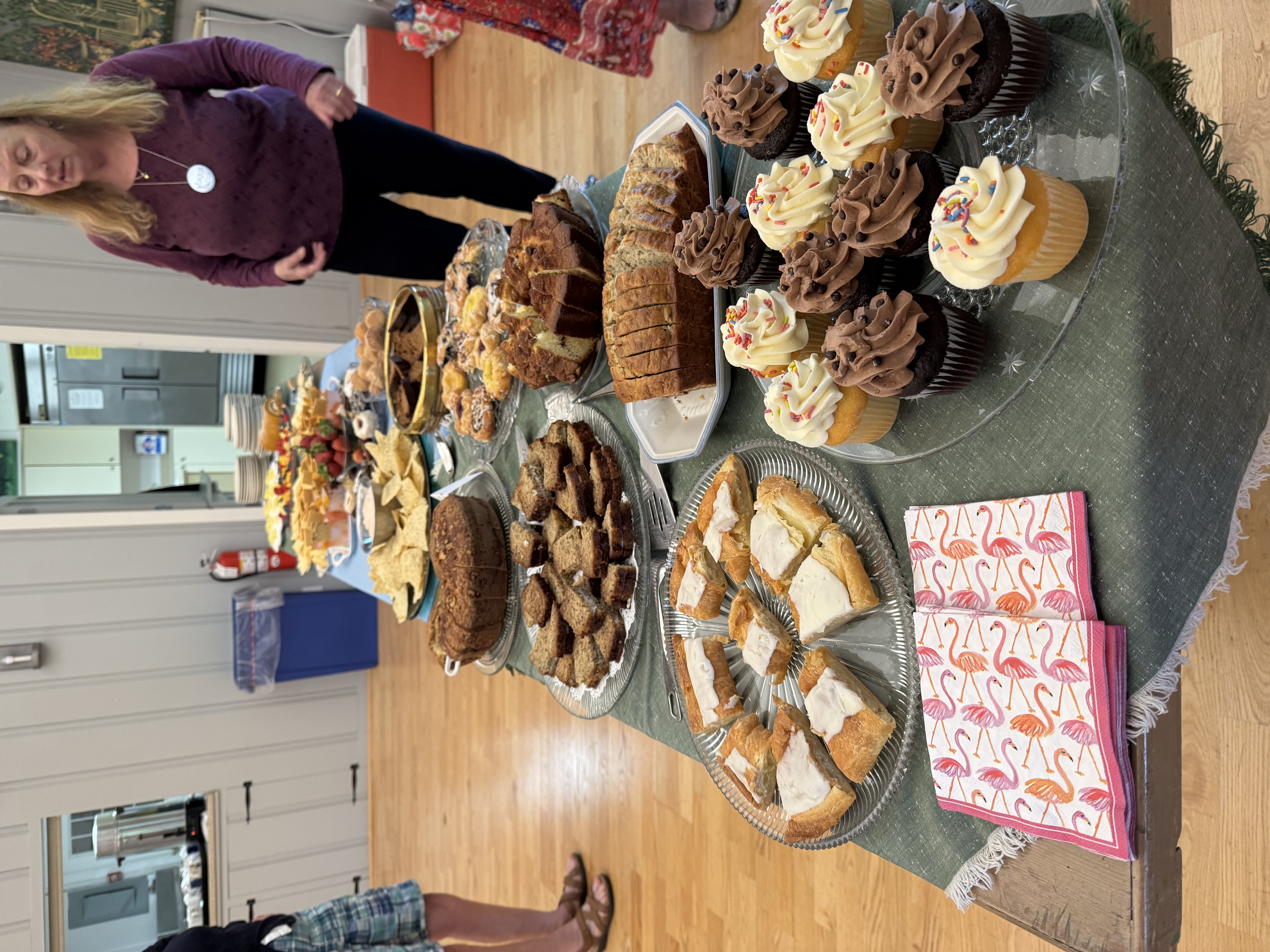 A woman pictured looking over a banquet table filled with plates of sweet treats like cupcakes, cookies, pastries and chips