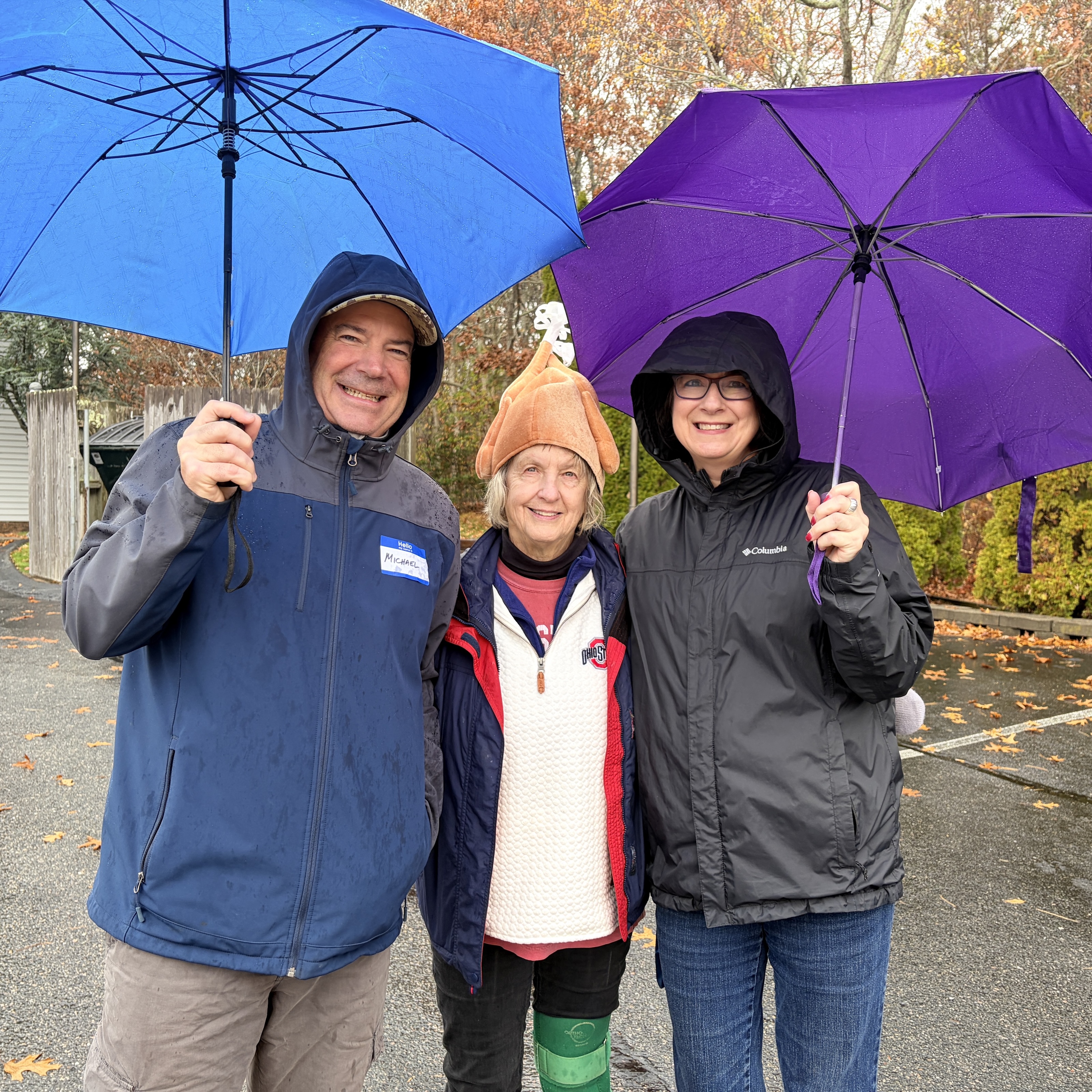 Three people with blue and purple umbrellas standing together in the rain in a parking lot